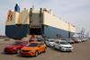 A large roll-on/roll-off cargo ship is docked at an industrial port, with multiple colour cars