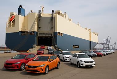 A large roll-on/roll-off cargo ship is docked at an industrial port, with multiple colour cars