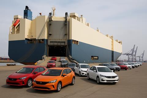 A large roll-on/roll-off cargo ship is docked at an industrial port, with multiple colour cars